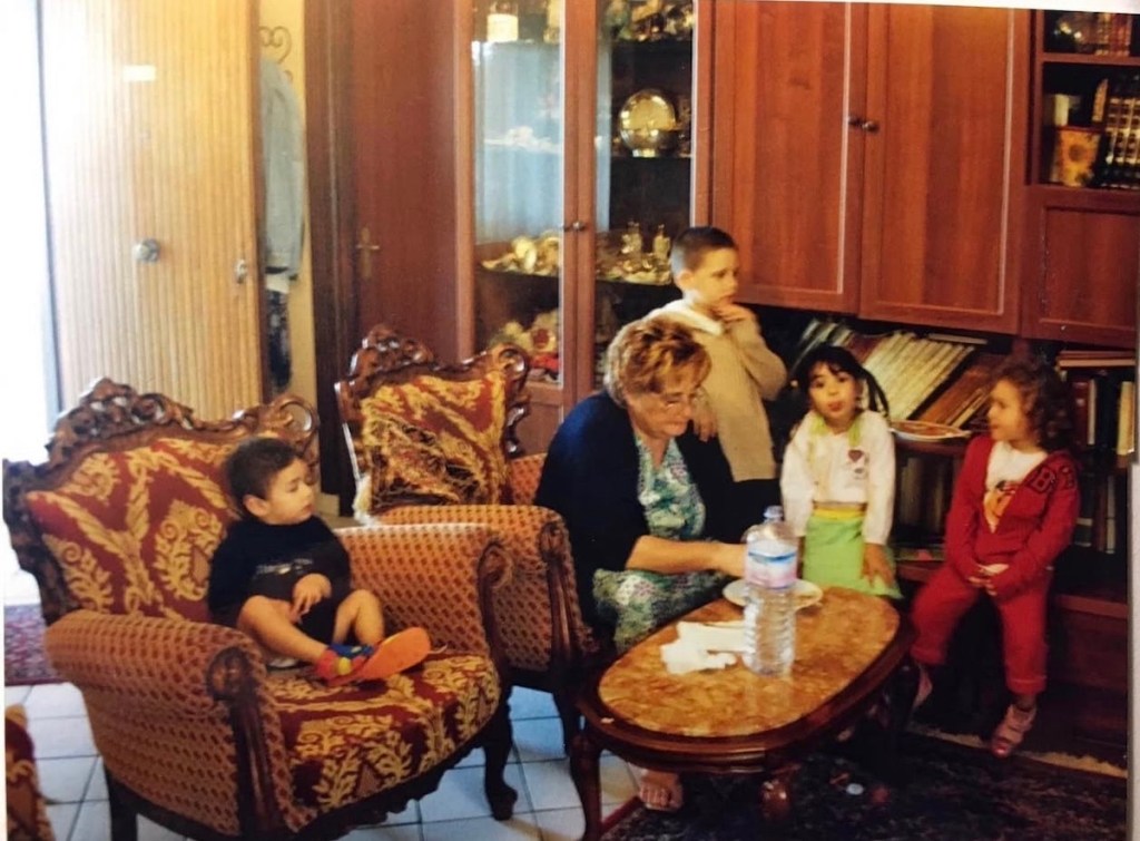 Un grupo de niños pequeños y un adulto se sientan en un salón con muebles ornamentados. Los niños parecen estar disfrutando de un momento juntos, mientras que el adulto, posiblemente una abuela, interactúa con ellos.