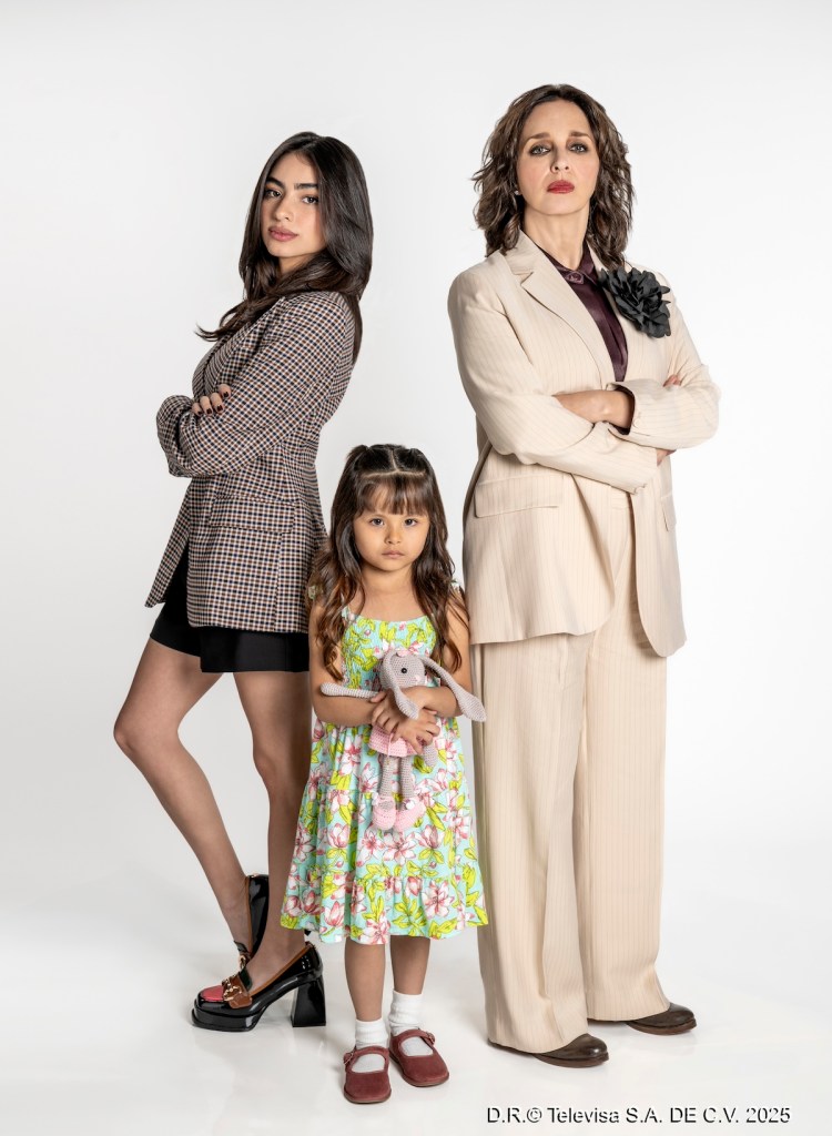 Tres mujeres de pie posan para la cámara, una de ellas sostiene un muñeco de peluche. La mujer a la derecha usa un traje claro y tiene una expresión seria, la mujer a la izquierda viste un conjunto más informal con un patrón de cuadros, y la niña en el centro lleva un vestido colorido.