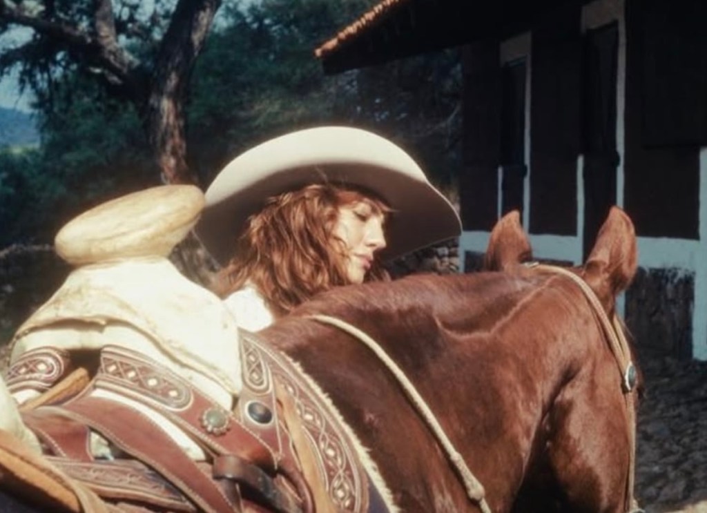Actriz montando un caballo, vista desde el lado, con un sombrero grande y fondo natural.