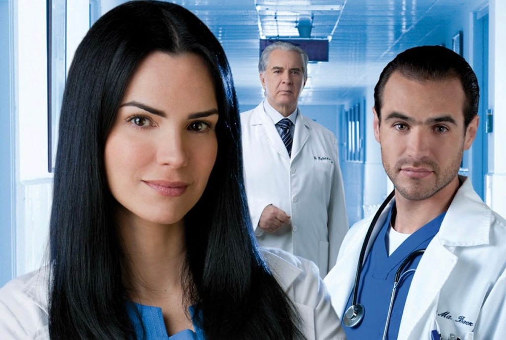 Tres médicos en una sala blanca, con un fondo de pasillo del hospital. La mujer, de cabello largo y negro, está al frente con una expresión seria. A su izquierda, un médico con un estetoscopio y a su derecha, otro médico de pie, con chaqueta blanca.