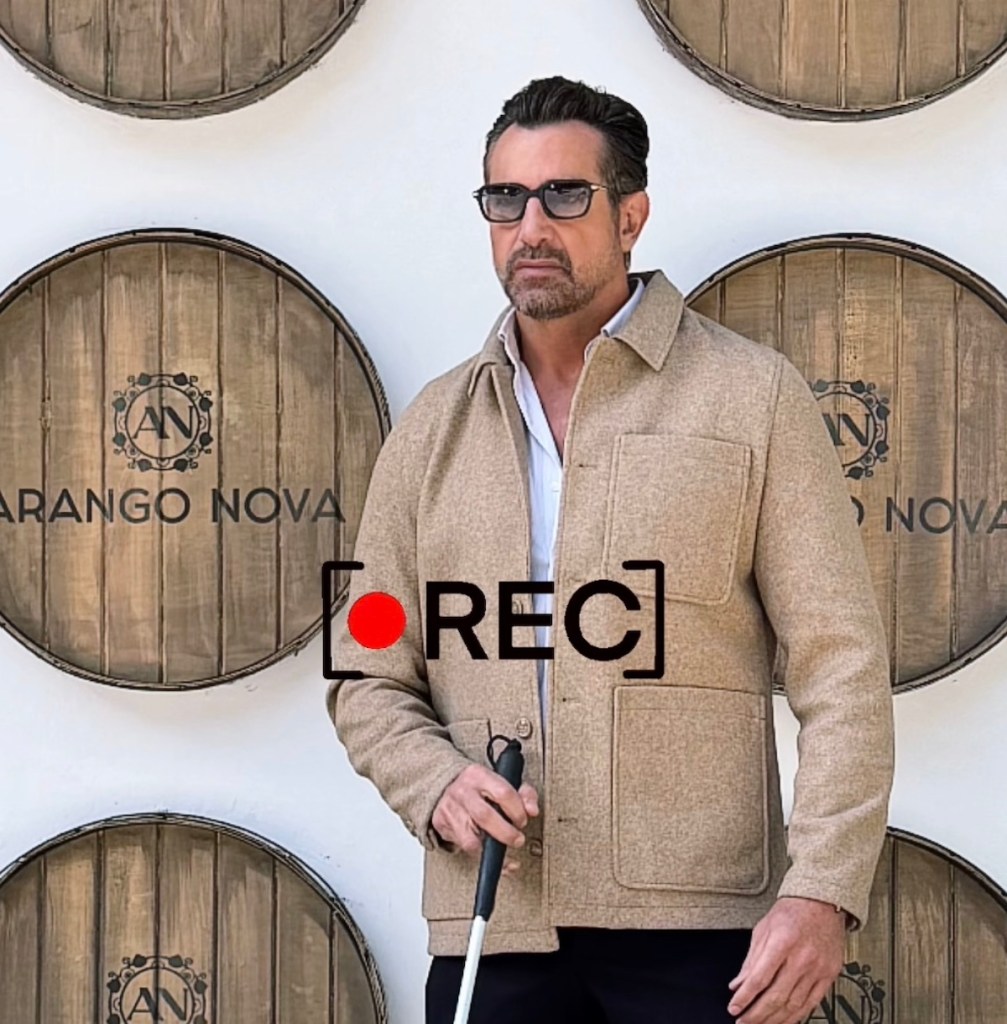 Hombre vistiendo una chaqueta beige y gafas oscuras, sosteniendo un bastón, de pie frente a barriles de madera con el logo de 'Arango Nova'.