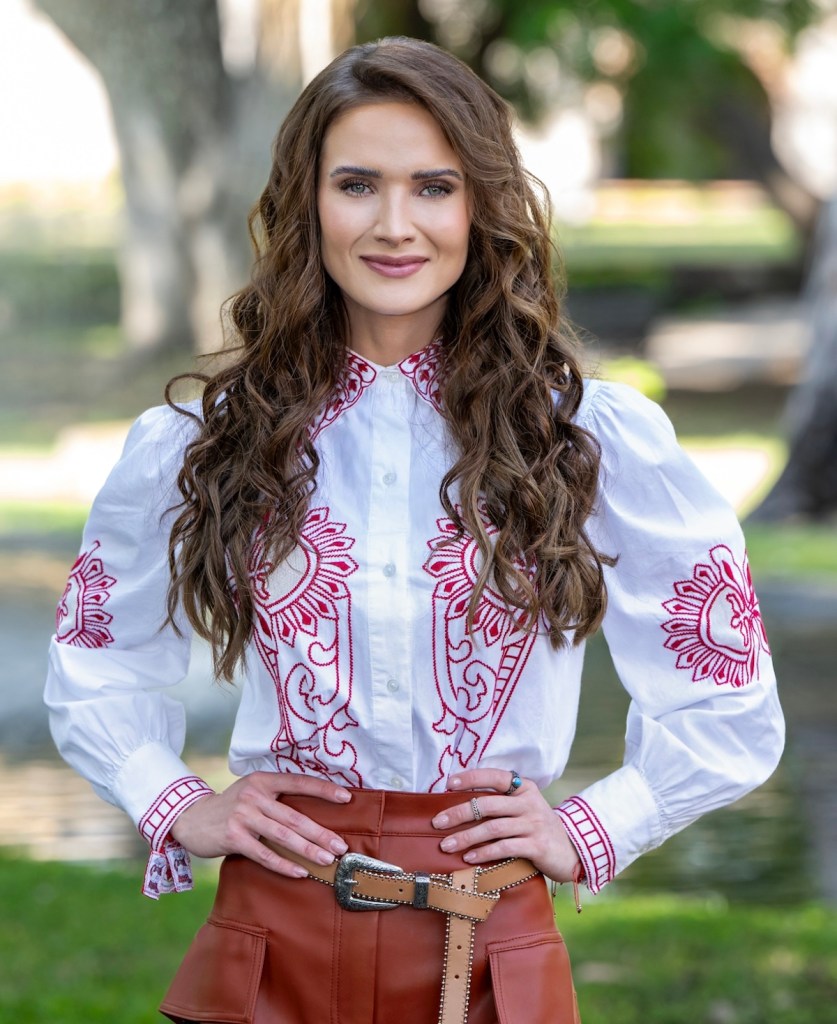 Mujer joven con cabello largo y rizado, sonriendo mientras lleva una camisa blanca con bordados rojos y una falda marrón, en un entorno al aire libre.