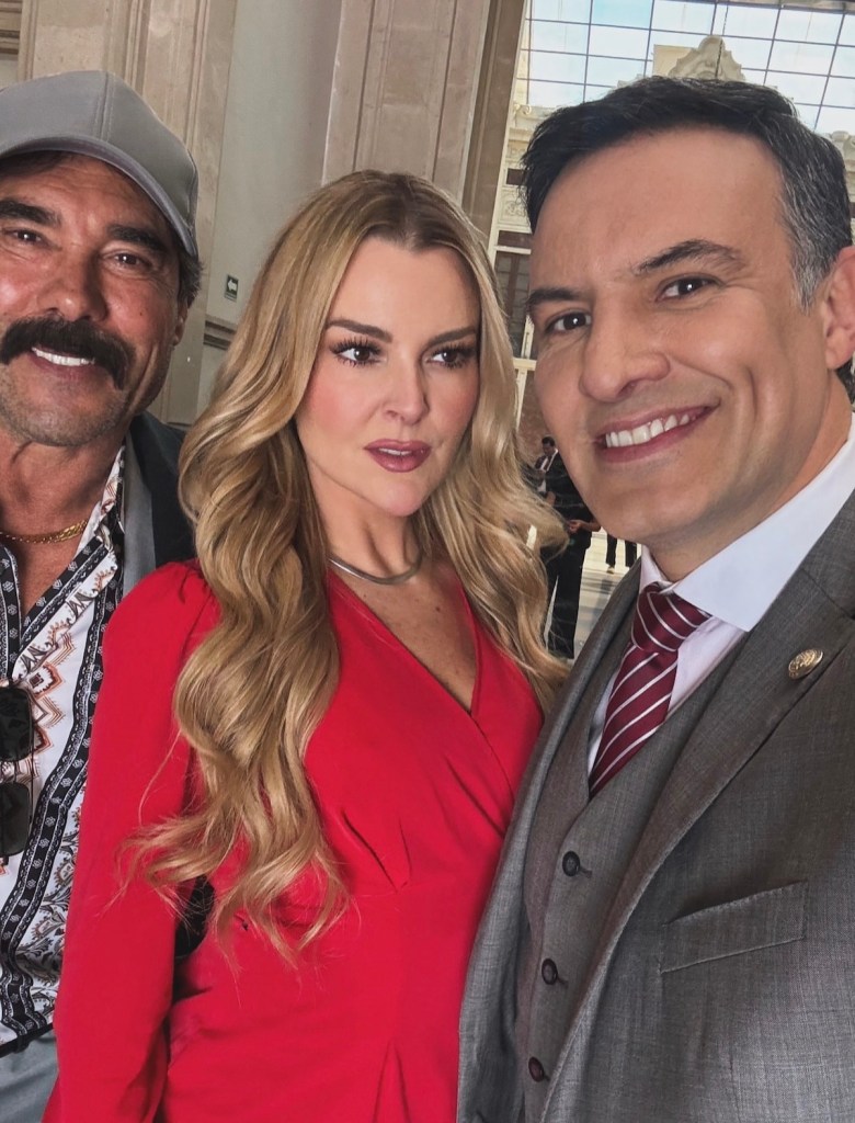 Marjorie de Sousa posando en un set de grabación con dos actores, ambos sonriendo. Ella lleva un vestido rojo, mientras que los otros dos actores están en atuendos formales.