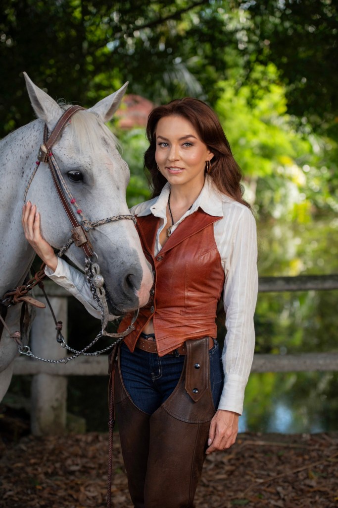 Mujer posando junto a un caballo blanco en un entorno natural, vestida con chaleco de cuero y pantalones de montar.