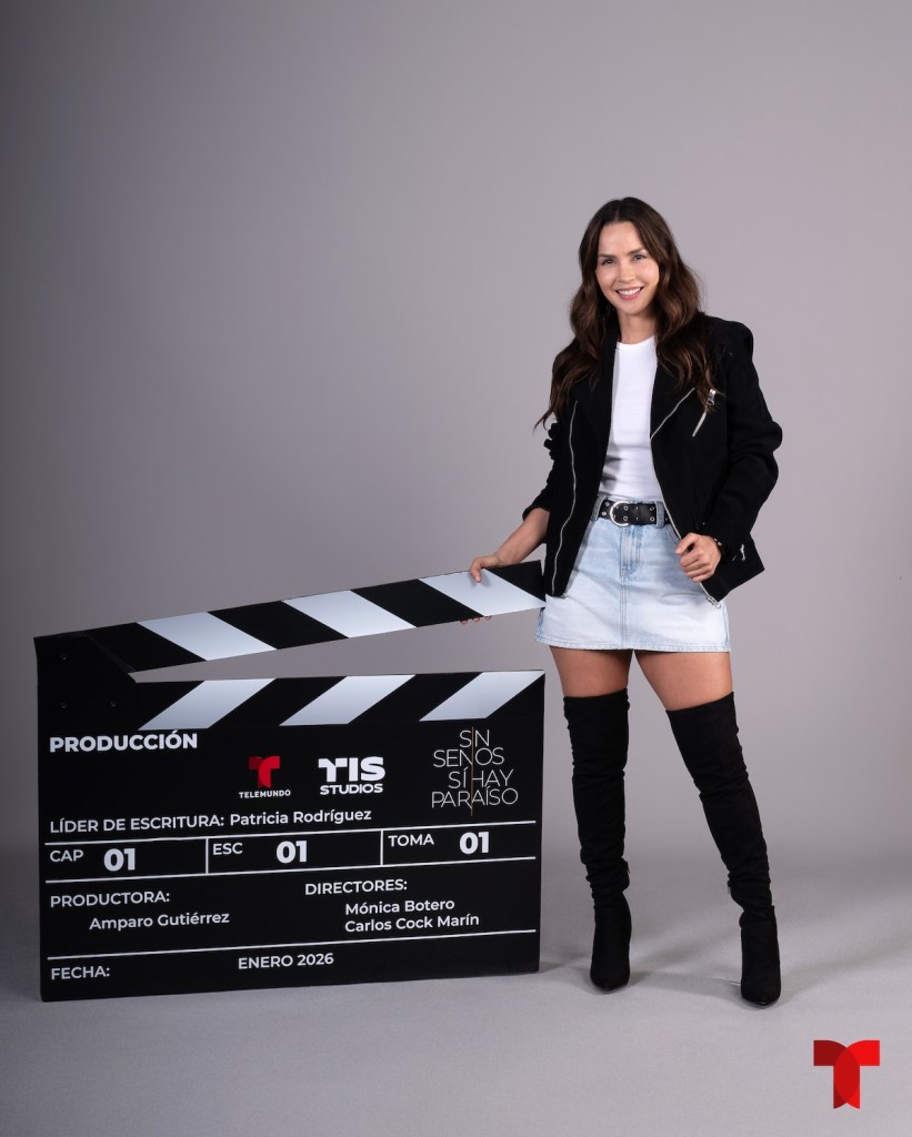 Mujer posando con una claqueta de producción que muestra información sobre un proyecto llamado 'Sin Senos Sí Hay Paraíso'. La mujer lleva una chaqueta negra y una falda de mezclilla, y está de pie con botas altas.