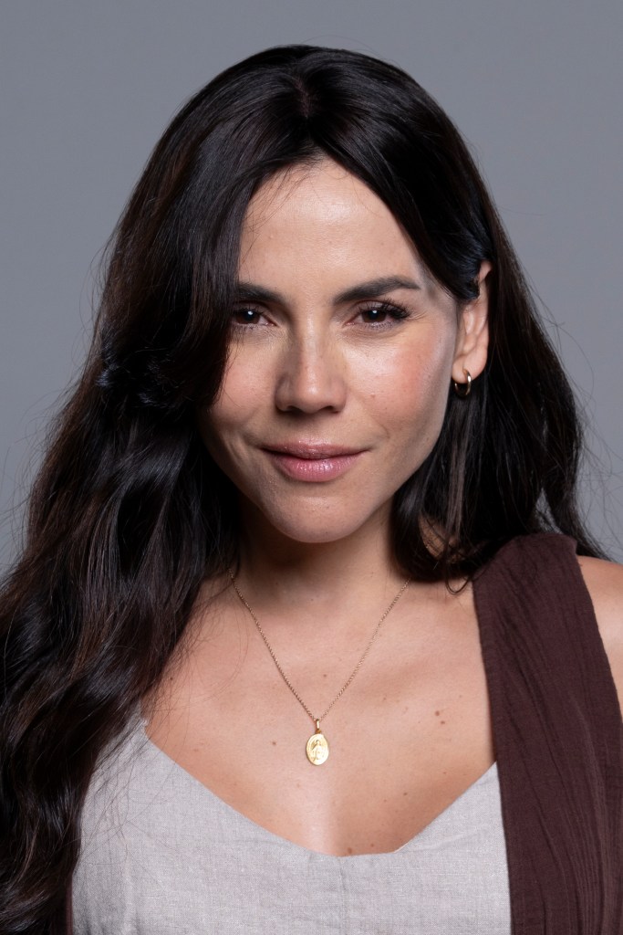 Retrato de una mujer con cabello largo y lacio, sonriendo suavemente, vestida con una blusa clara y un chaleco oscuro. Ella lleva un collar dorado.