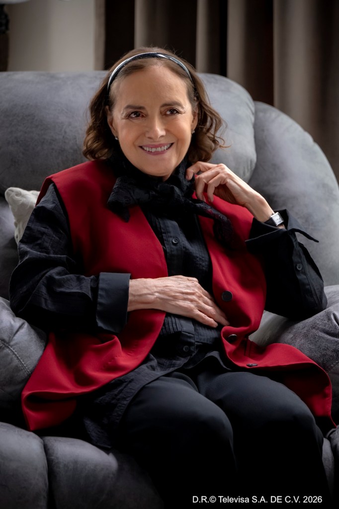 Mujer sonriendo sentada en un sofá, vistiendo un chaleco rojo y camisa negra con un lazo negro.