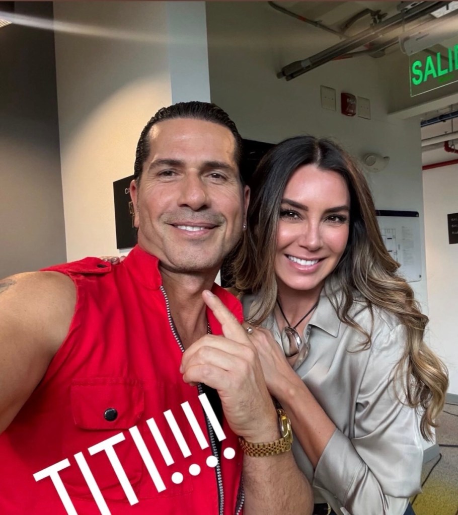 Selfie de dos personas sonriendo, un hombre con camiseta roja y una mujer con blusa gris, ambos mostrando una mano cerca de la cámara.