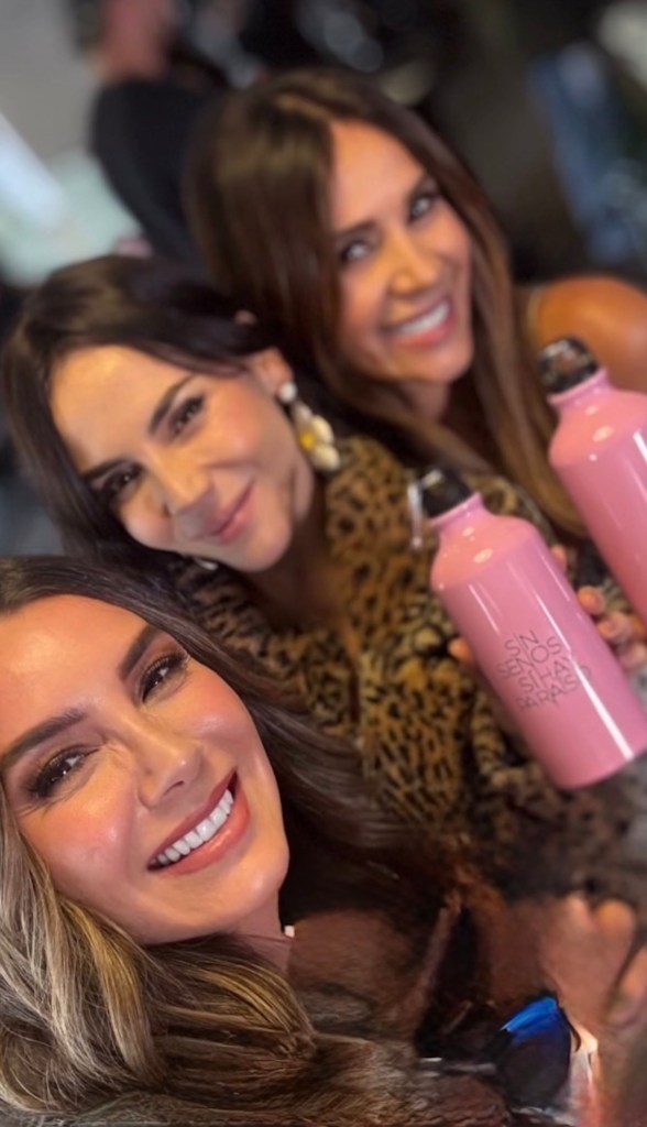 Tres mujeres sonrientes posando juntas y sosteniendo botellas de agua rosas, en un ambiente alegre.