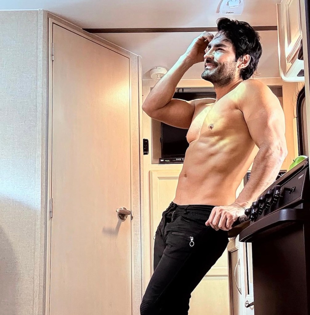 Hombre musculoso posando sin camiseta en un entorno interior, sonriendo mientras se acaricia la cabeza, vistiendo pantalones oscuros.
