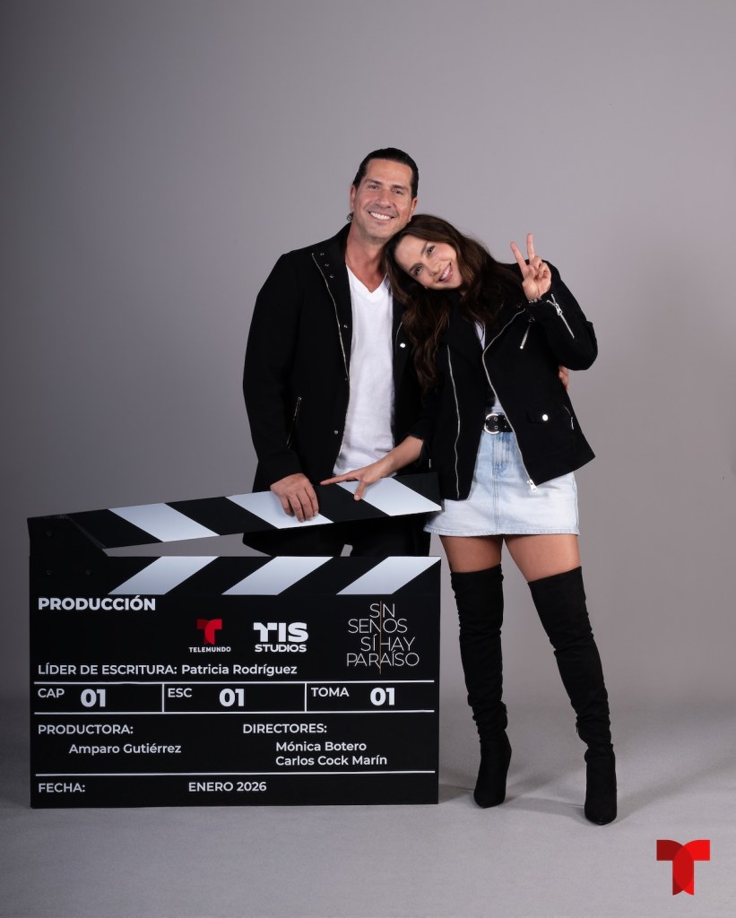 Dos personas posando con un claquetazo de cine, el hombre sonríe mientras la mujer hace una señal de paz. Ambos visten chaquetas negras y aparecen en un fondo gris.