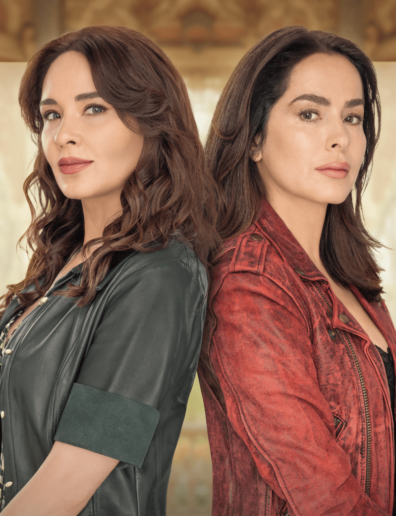 Dos mujeres de cabello castaño y lacio, una vestida con una chaqueta de cuero negra y la otra con una chaqueta de cuero roja, posando de espaldas una a la otra en un fondo difuminado.