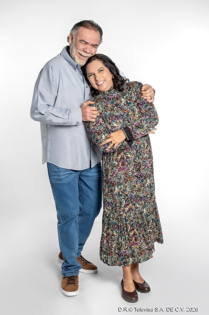 Pareja sonriente posando juntos, el hombre lleva una camisa clara y jeans, mientras que la mujer viste un vestido largo de estampado colorido.