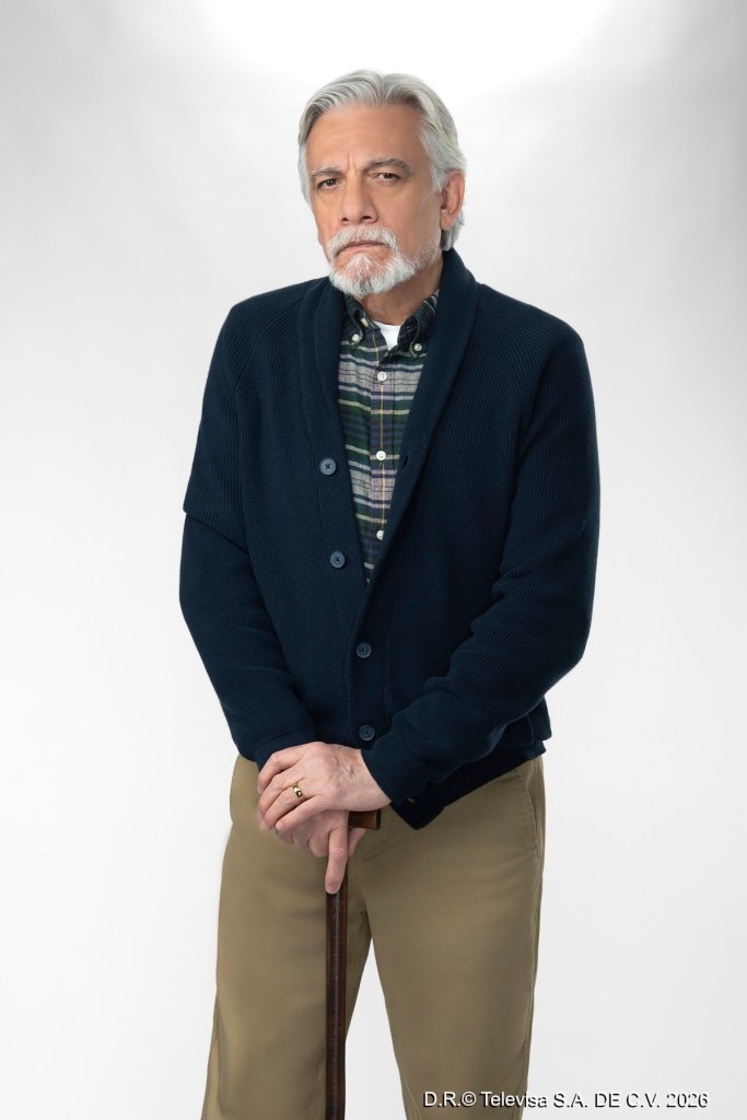 Hombre mayor con cabello gris y barba, vestido con un suéter azul marino y una camisa de cuadros. Sostiene un bastón y mira a la cámara con una expresión seria.