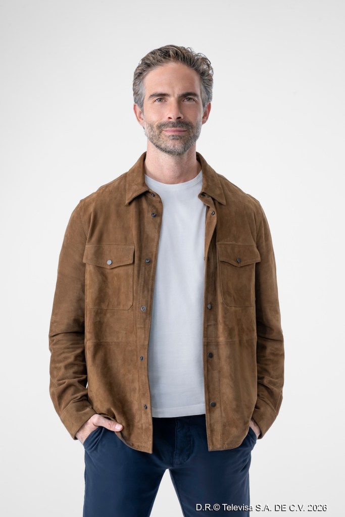 Hombre sonriendo con chaleco de ante marrón y camiseta blanca, de pie contra un fondo blanco.
