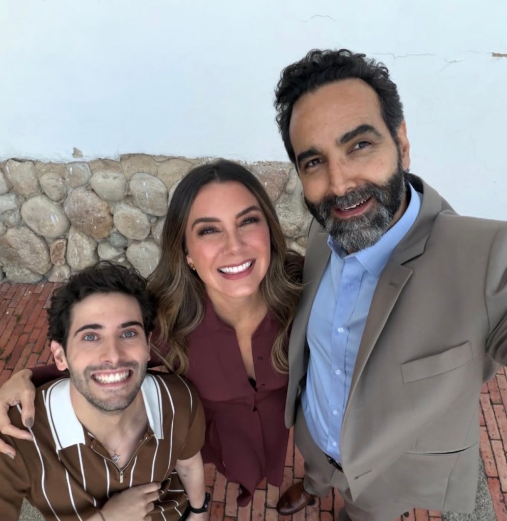 Tres personas sonriendo en una foto grupal, con un fondo de pared de piedra y suelo de ladrillos. Dos hombres y una mujer, la mujer lleva un vestido marrón y los hombres una camisa de rayas y un traje claro.
