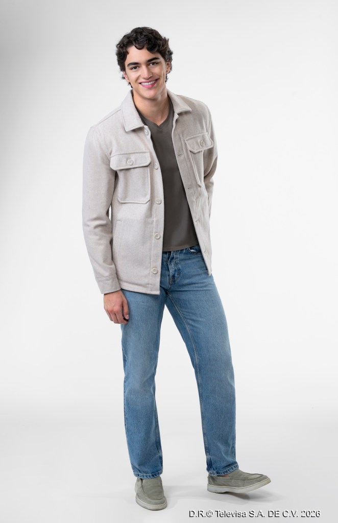 Joven sonriente de pie, vistiendo una chaqueta clara, camiseta gris y jeans azules, sobre un fondo blanco.