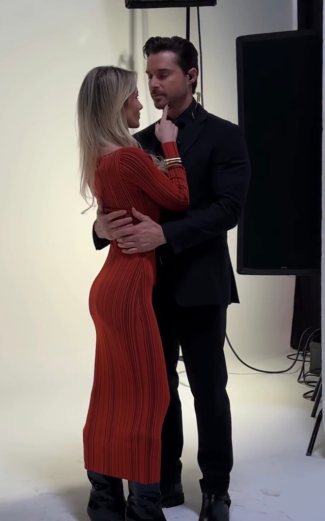 Pareja abrazada en un estudio fotográfico, la mujer lleva un vestido rojo ajustado y el hombre un traje negro. Se miran con complicidad mientras ella toca suavemente su rostro.
