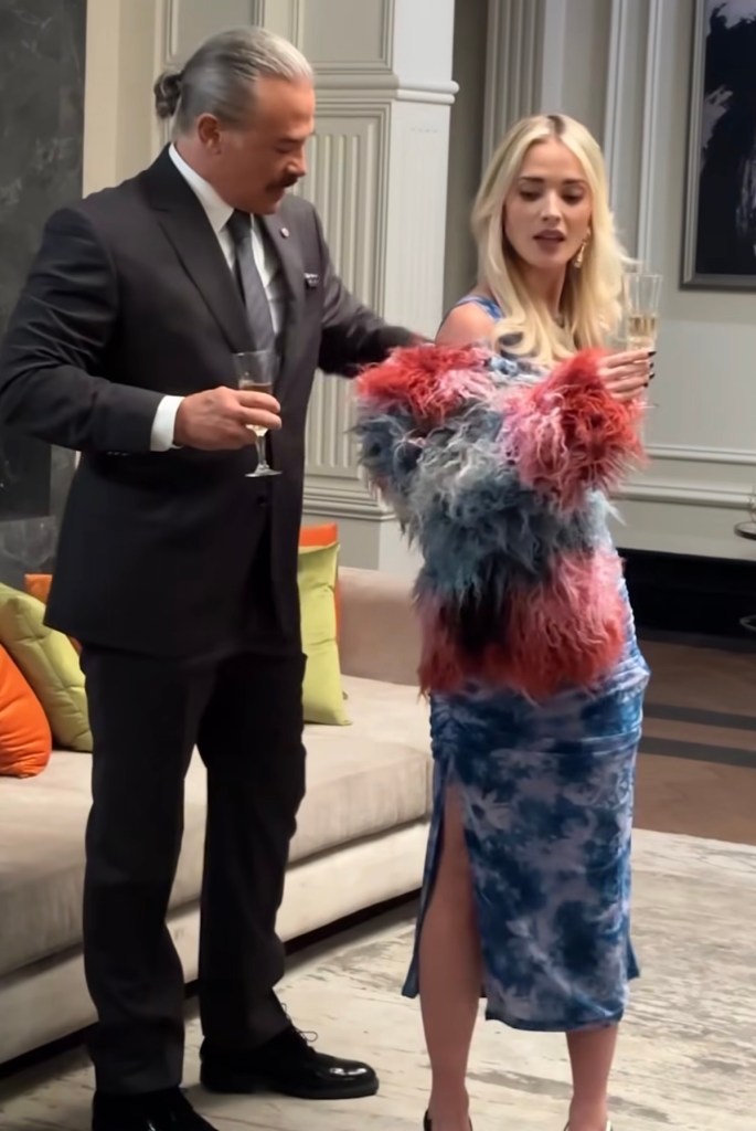 Un hombre vestido de traje sostiene una copa de champán mientras observa a una mujer con un vestido azul y un abrigo de pelo colorido. Están en un ambiente elegante.