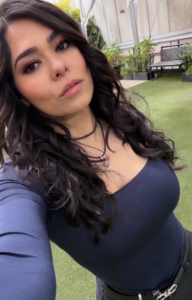 Autorretrato de una mujer con cabello largo y rizado, vestida con una blusa de mangas largas oscura, en un entorno al aire libre con plantas.