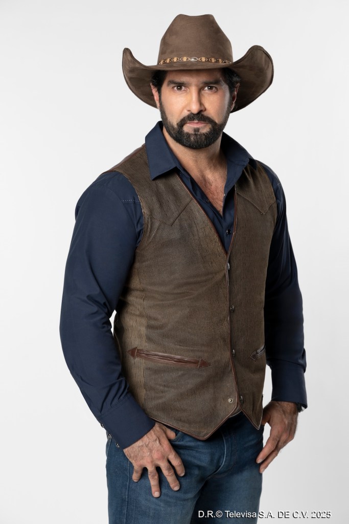 Hombre con barba y cabello oscuro, vistiendo un sombrero marrón y un chaleco de tonos oscuros, posando de forma seria frente a un fondo blanco.