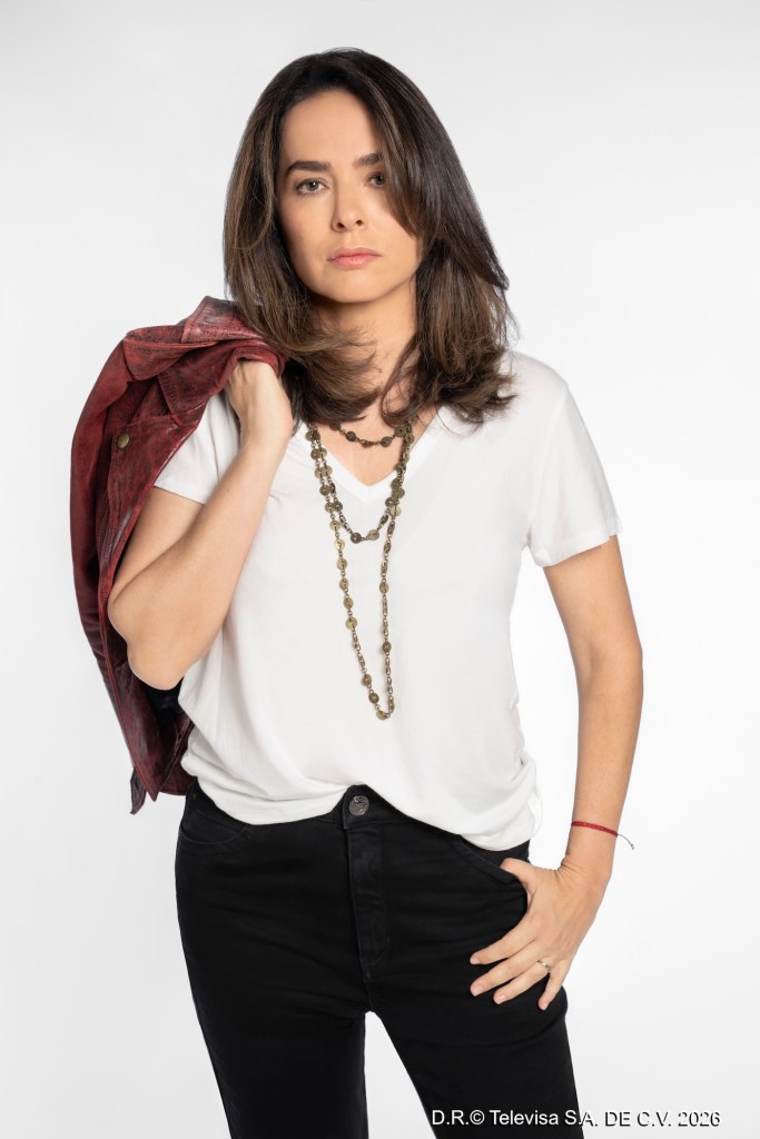 Mujer con cabello largo y suelto, viste una camiseta blanca y jeans negros, con una chaqueta roja sobre el hombro. Lleva un collar y muestra una expresión seria.