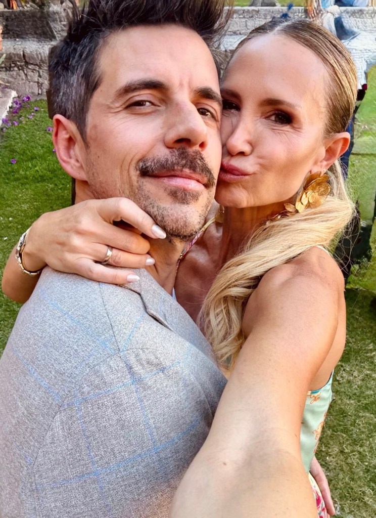 Pareja sonriente tomando un selfie en un ambiente al aire libre con césped y flores de fondo.