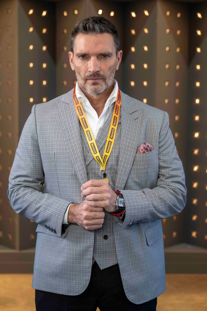 Hombre vestido con un traje claro y corbata, sosteniendo un medallero con cintas amarillas en un fondo decorativo.
