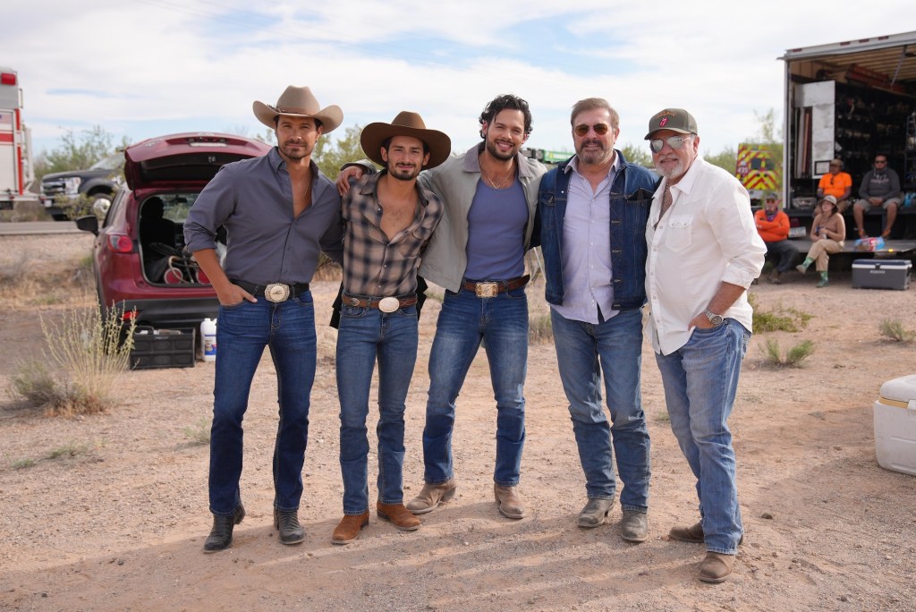 Grupo de cinco hombres posando juntos al aire libre, vestidos con ropa informal y sombreros, en un entorno desértico con vehículos y equipo de fondo.