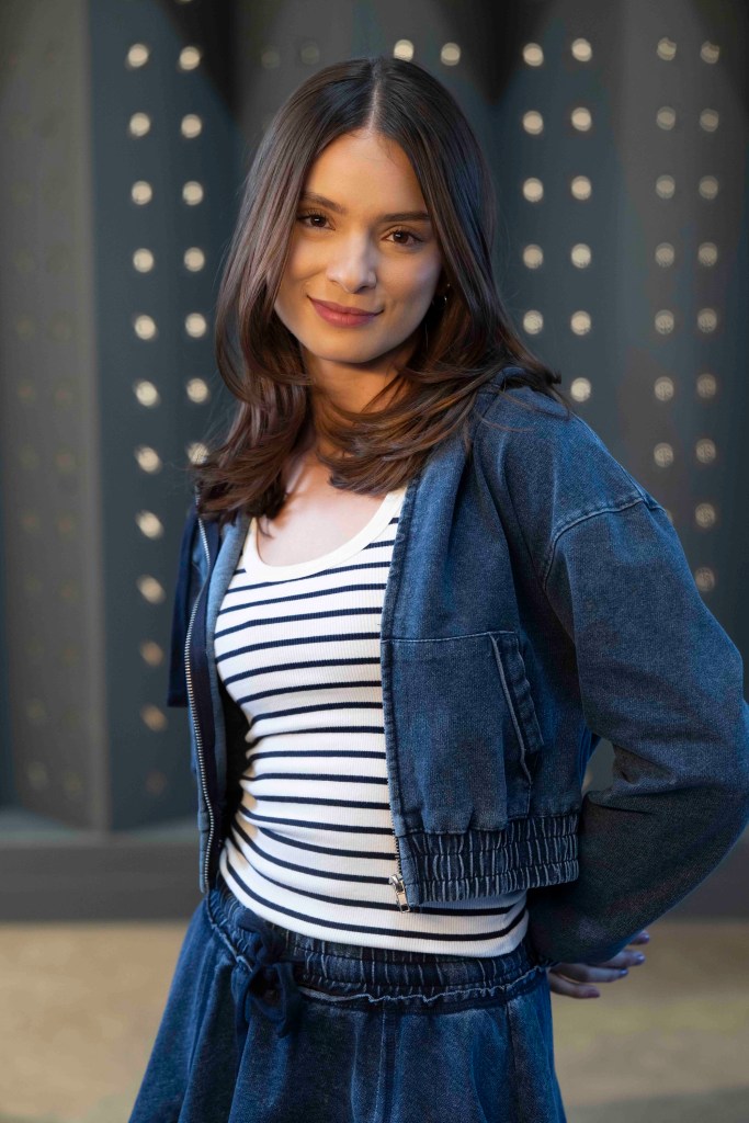 Mujer sonriendo con un atuendo de mezclilla, compuesto por una chaqueta corta y un pantalón de tiro alto, de pie frente a un fondo con luces brillantes.