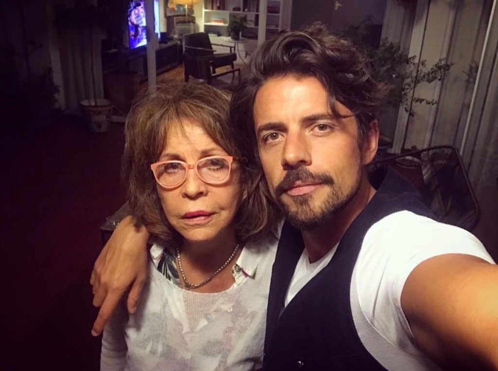 Dos personas posan para un selfie, una mujer con gafas y una expresión seria, y un hombre con barba sonriente, en un ambiente interior acogedor.